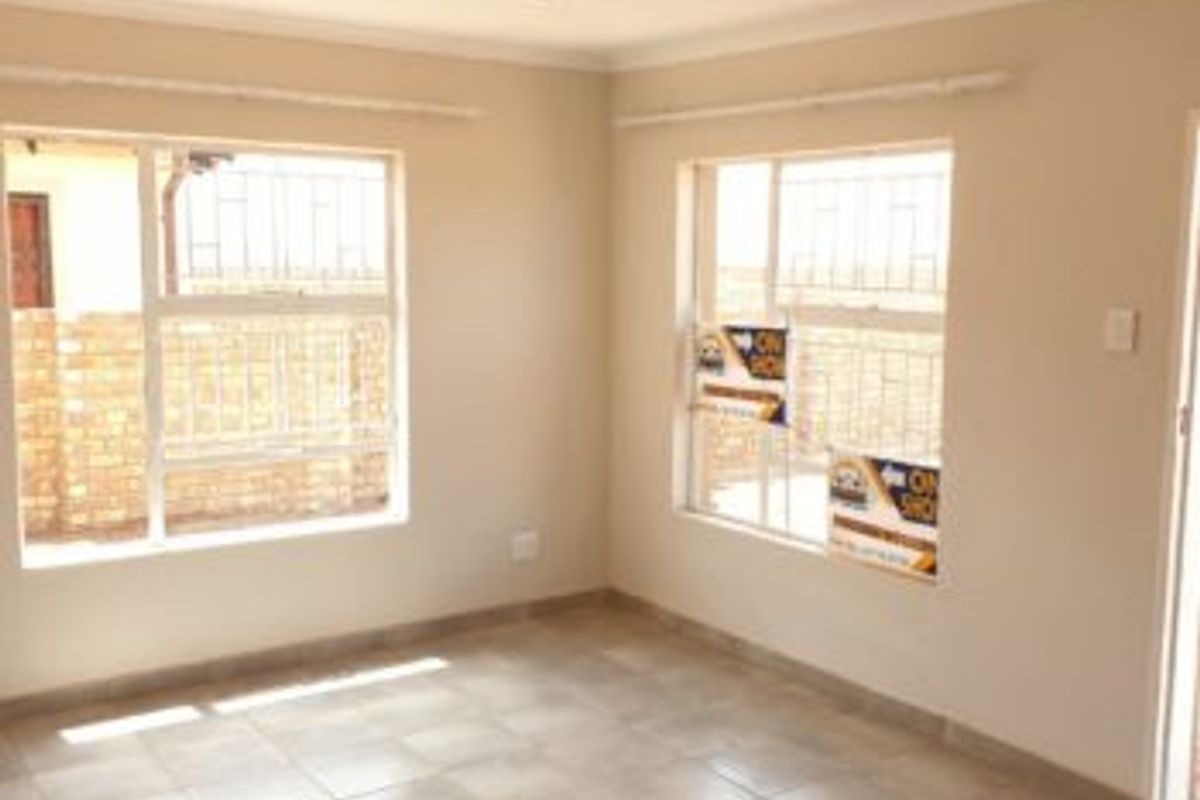 A bright, empty living room featuring neutral tiled flooring, recessed ceiling lighting, and large windows with security bars.