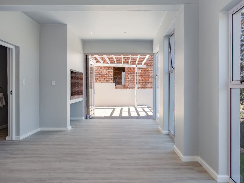 Bright open-plan living area featuring light wood-look flooring, a built-in indoor braai, and seamless access to an outdoor patio area.