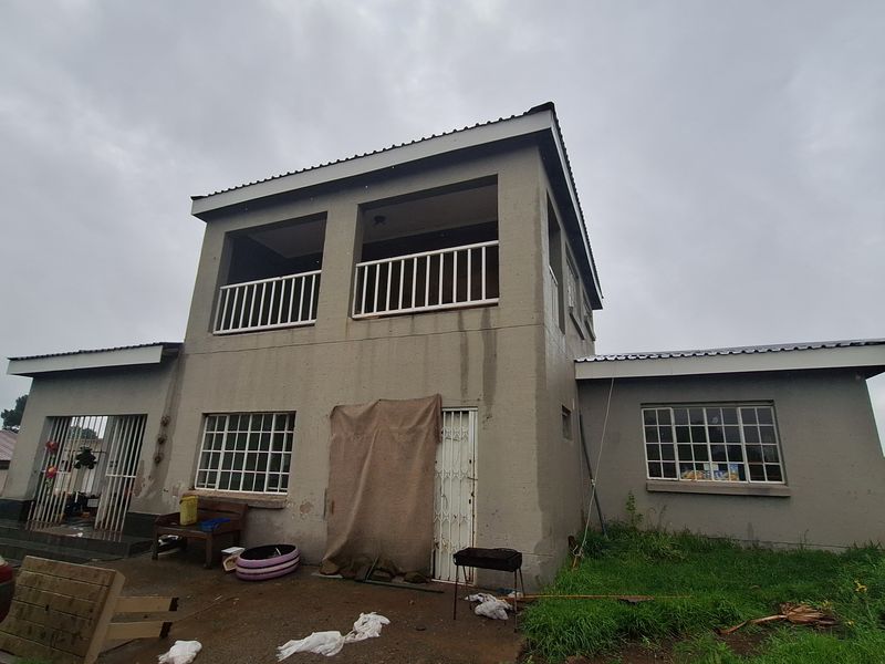 Two-story residential property featuring a balcony, metal security gates, and windows with burglar bars, set on a plot with a concrete patio area.