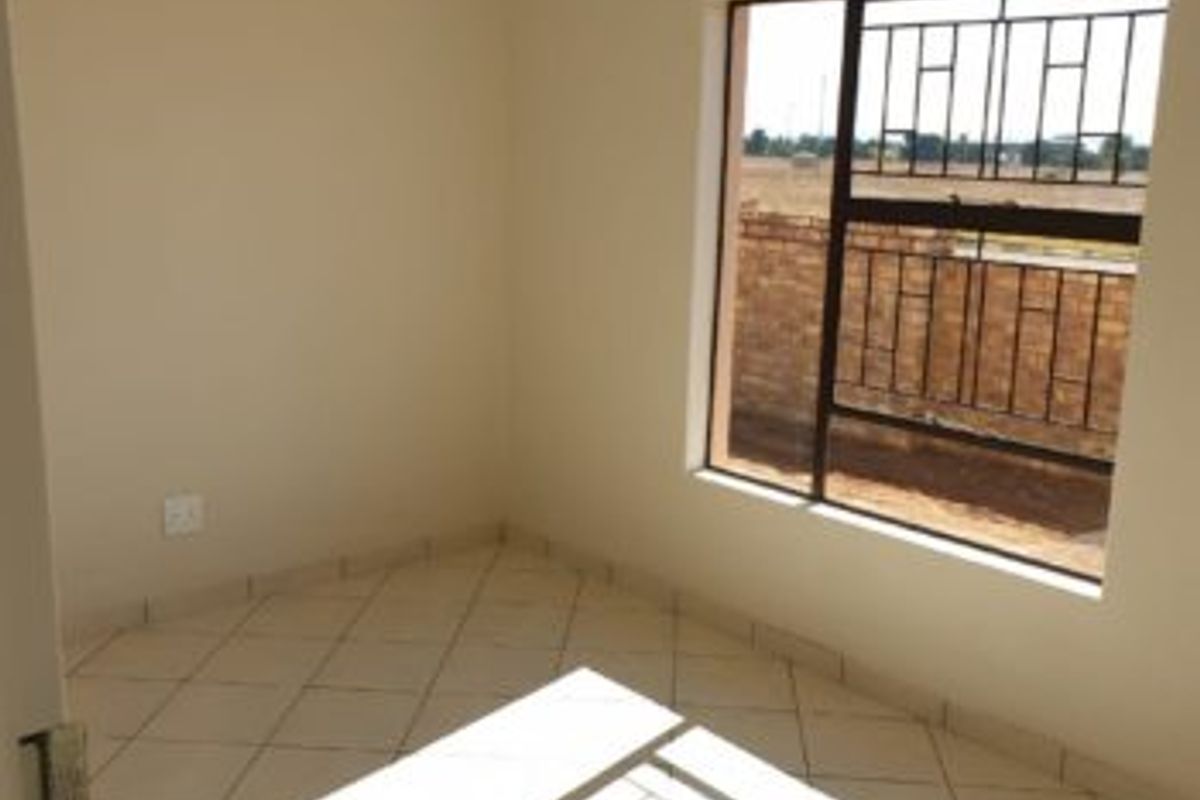 A bright, empty bedroom featuring neutral wall paint, tiled flooring, and a window fitted with security bars.