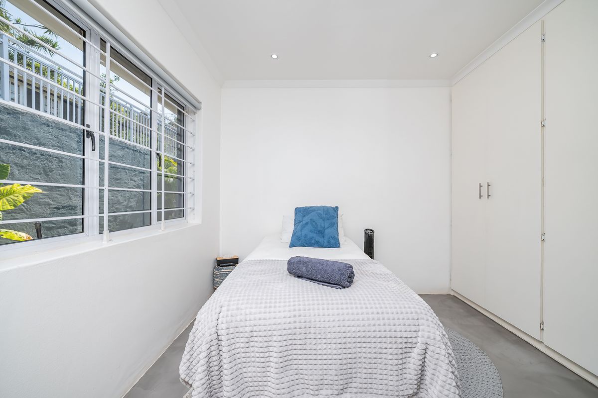 A bright bedroom featuring built-in wardrobes, recessed ceiling lighting, and a large window with security bars overlooking an exterior retaining wall.