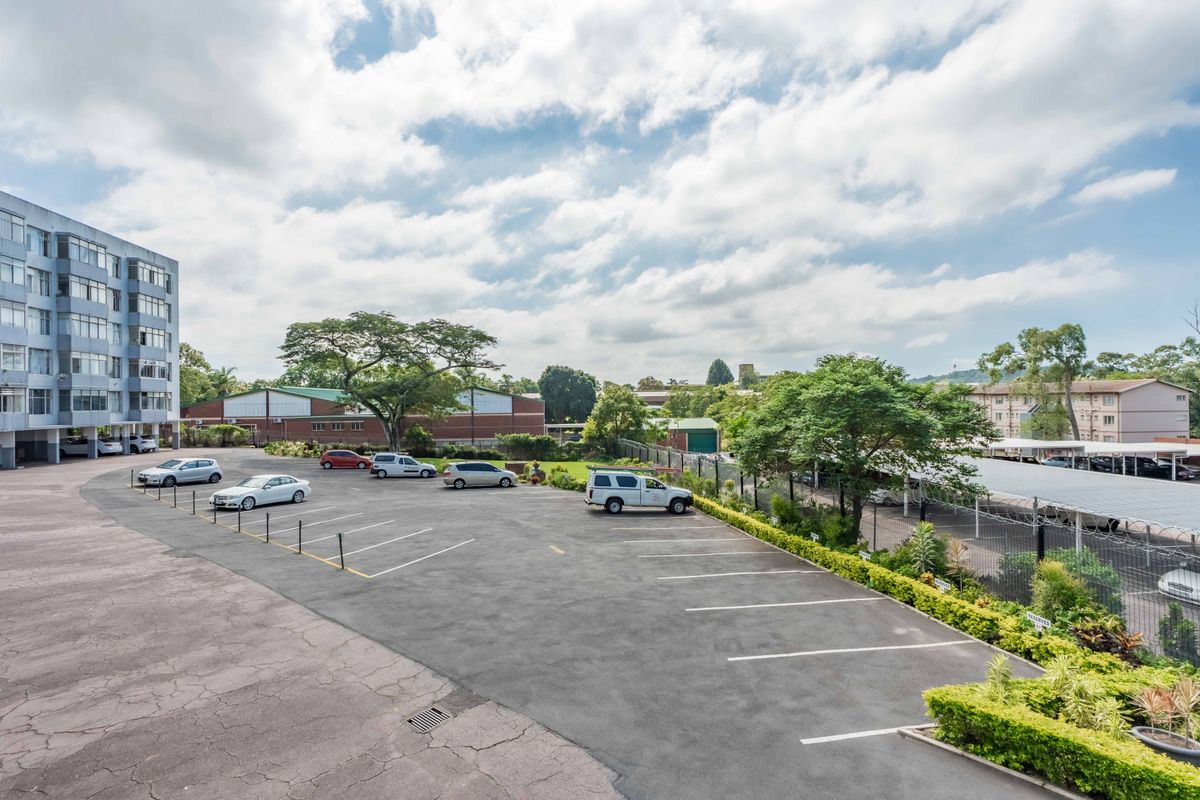 An elevated view of the apartment complex parking area featuring asphalt paving, marked bays, perimeter fencing, and landscaped garden borders.