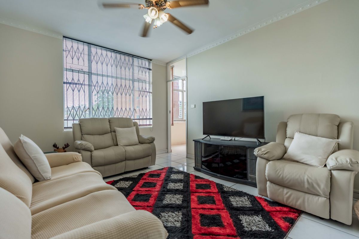 A comfortable living room featuring tiled flooring, a ceiling fan, and a large window with security bars, providing access to an adjacent balcony.