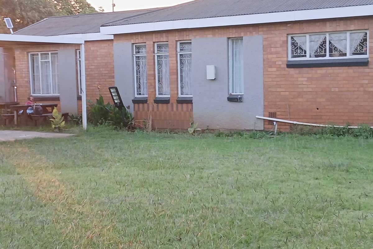 Rear view of a single-story brick house featuring a corrugated metal roof, security-barred windows, and a small covered patio area overlooking the garden.