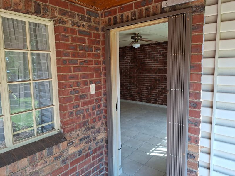 A covered patio area featuring exposed brickwork, tiled flooring, and a sliding security gate leading into an interior living space with a ceiling fan.