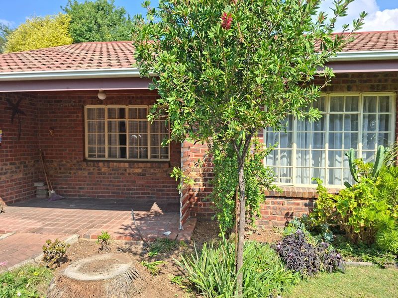 A rear view of a brick retirement village unit featuring a covered patio, tiled roof, and established garden area.