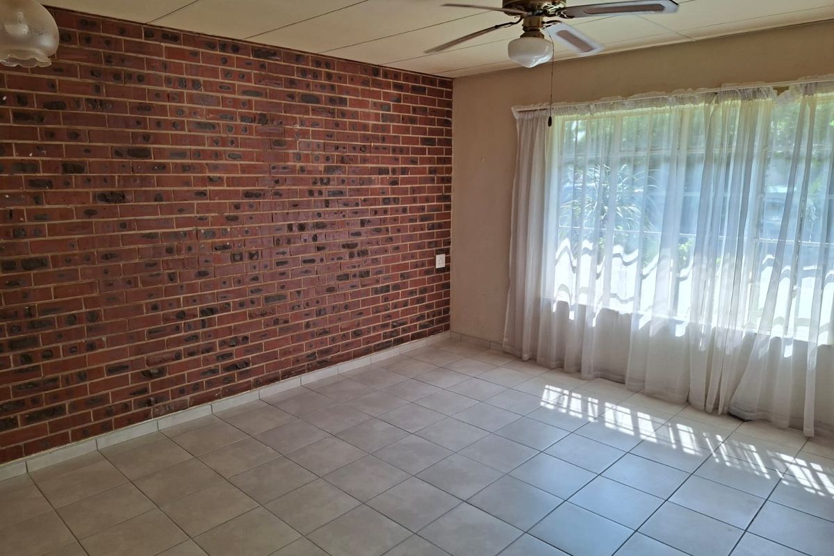 A bright, empty living room featuring a prominent exposed brick wall, neutral tiled flooring, and a ceiling fan, offering a blank canvas for personalization.