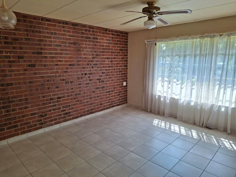 A bright, empty living room featuring a prominent exposed brick wall, neutral tiled flooring, and a ceiling fan, offering a blank canvas for personalization.