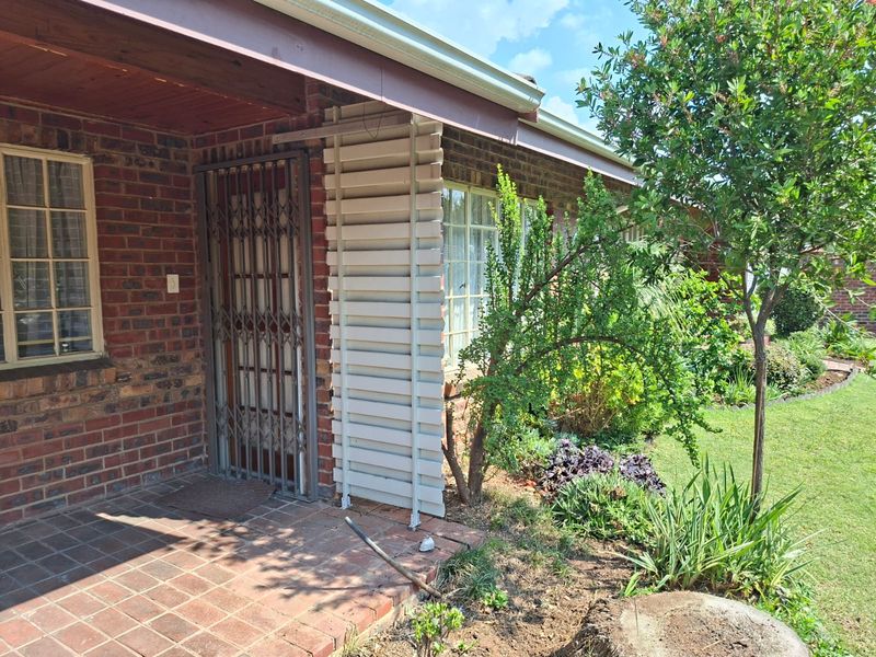 A covered brick patio area featuring a security gate, tiled flooring, and a louvered privacy screen, overlooking a landscaped garden.