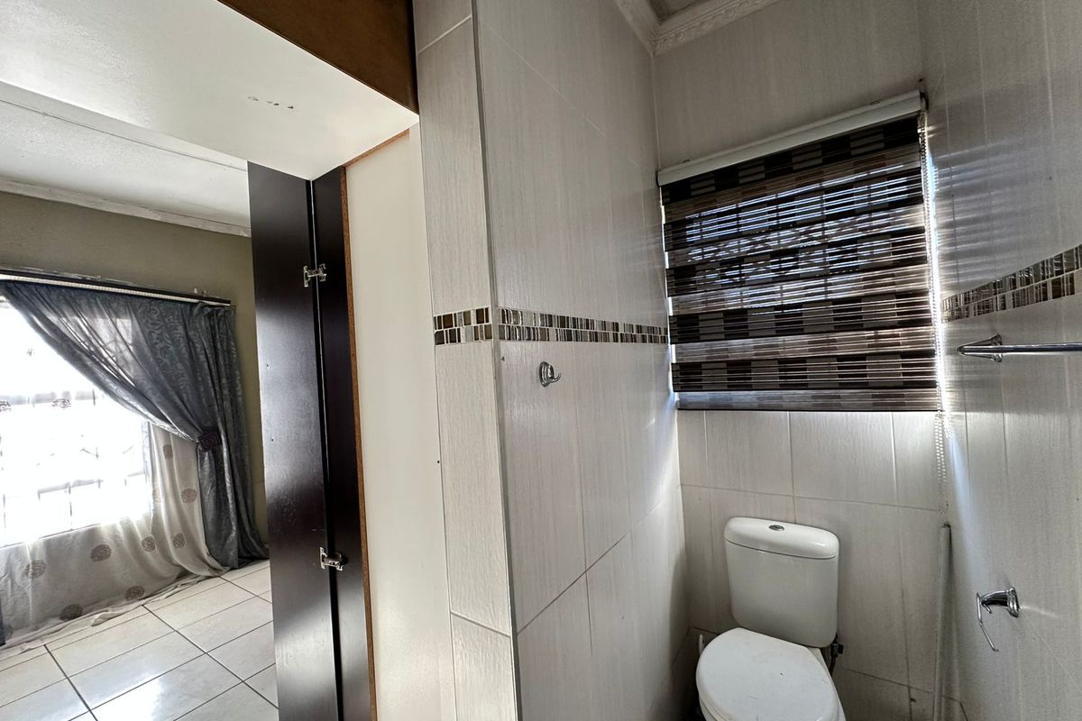 A functional bathroom featuring tiled walls, a standard toilet, and a window with a fitted blind, adjacent to a living area.