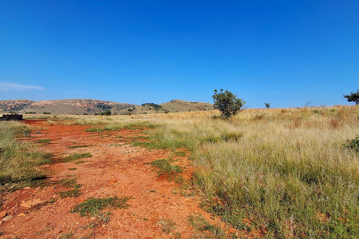 A wide-angle view of vacant residential land featuring natural grass, red soil, and distant hilly terrain under a clear blue sky.