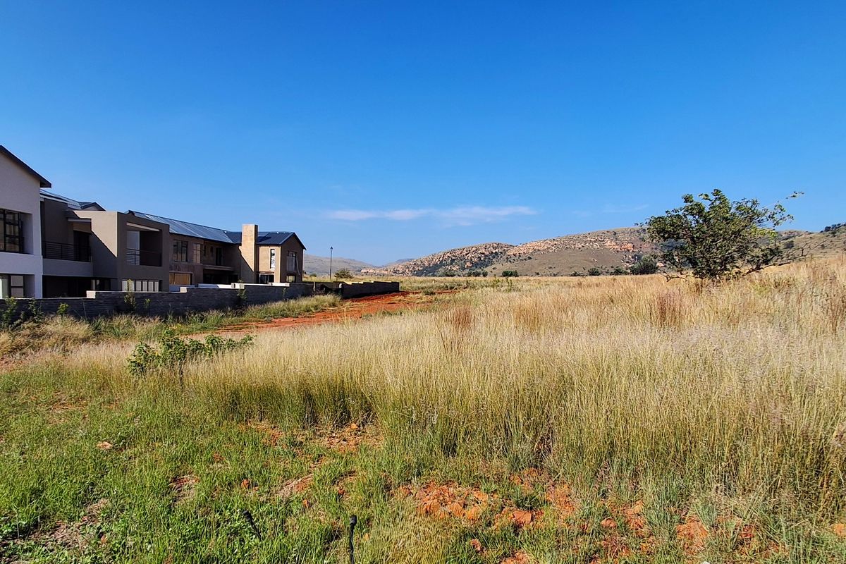A view of an undeveloped residential plot featuring natural grassland with adjacent modern housing and scenic mountain views in the background.