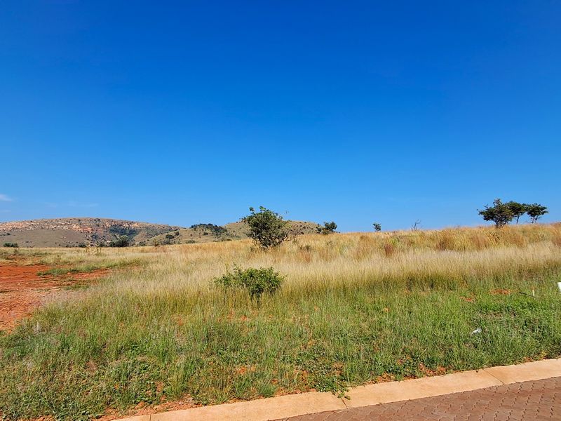 A view of a vacant residential plot featuring natural grass, scattered shrubs, and distant hilly landscape under a clear blue sky.