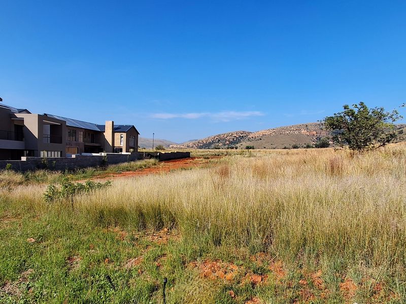 A view of an undeveloped residential plot featuring natural grassland with adjacent modern housing and scenic mountain views in the background.