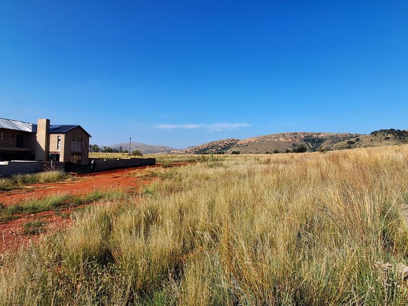 A view of a large vacant residential plot featuring natural grassland, a boundary wall, and a partially constructed dwelling in the background.