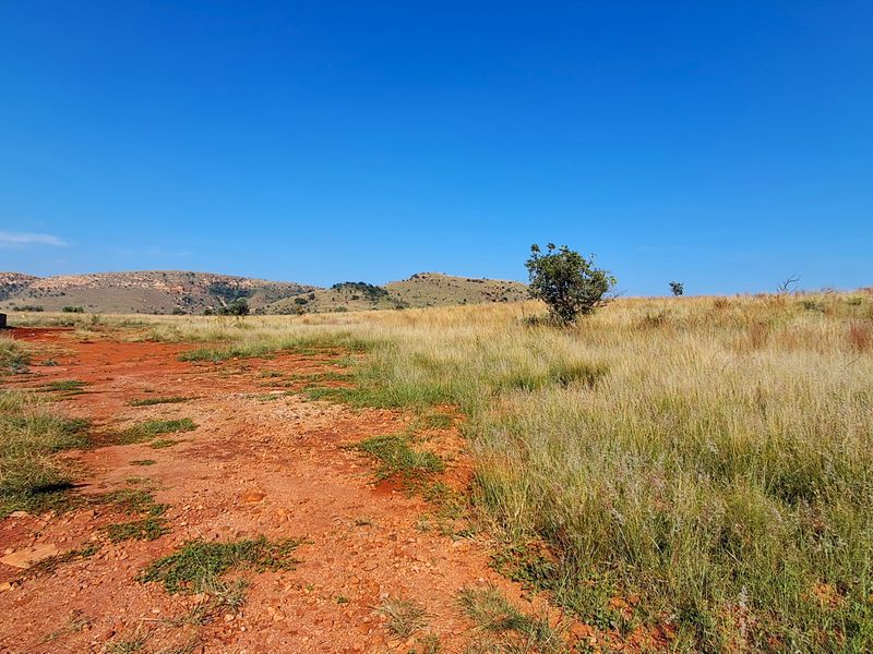 A wide-angle view of vacant residential land featuring natural grass, red soil, and distant hilly terrain under a clear blue sky.
