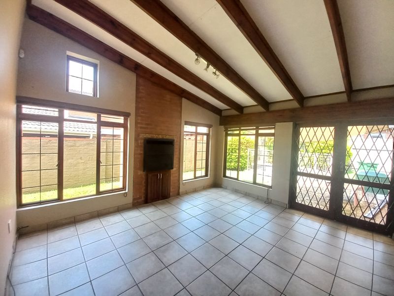 Spacious living room featuring exposed timber ceiling beams, a brick fireplace, tiled flooring, and large windows providing ample natural light.