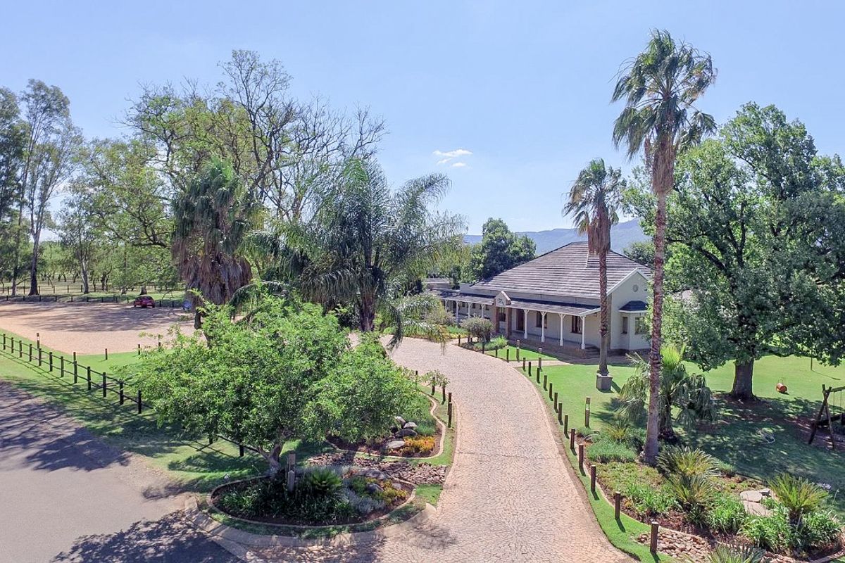 Aerial view of a residential property featuring a long paved driveway, expansive landscaped gardens, mature trees, and a main dwelling with a covered veranda.