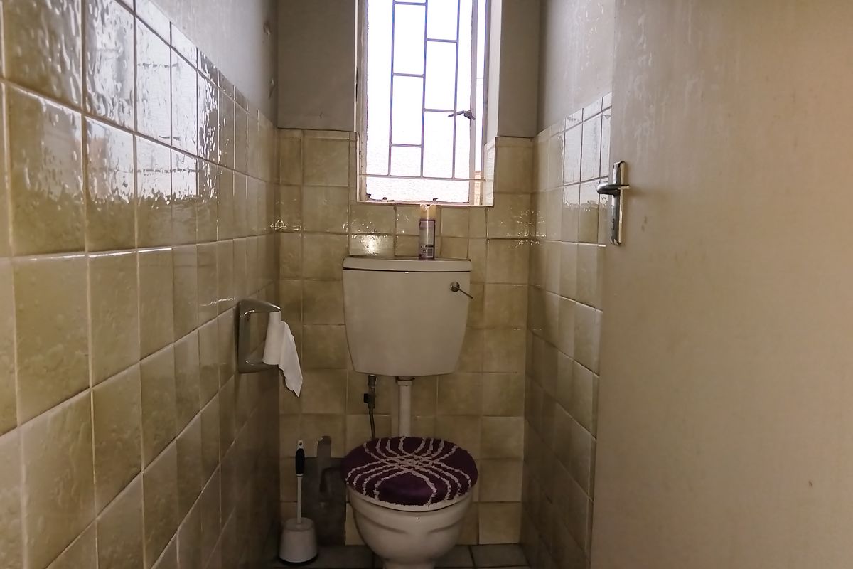 A compact, dated bathroom featuring ceramic wall and floor tiles, a standard toilet, and a window with security bars.
