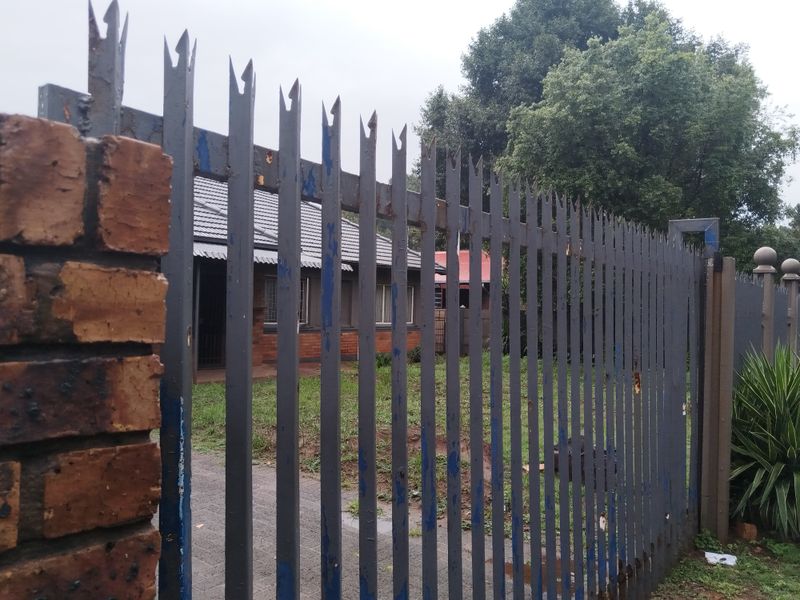 Front view of the property featuring a metal spike security fence, brick pillar, and paved driveway leading to the house entrance.