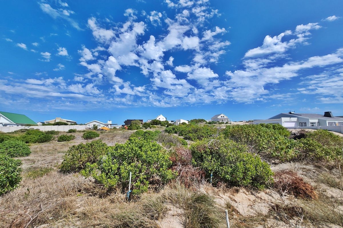 A view of a vacant residential plot featuring natural coastal vegetation and sandy terrain, surrounded by established residential properties.