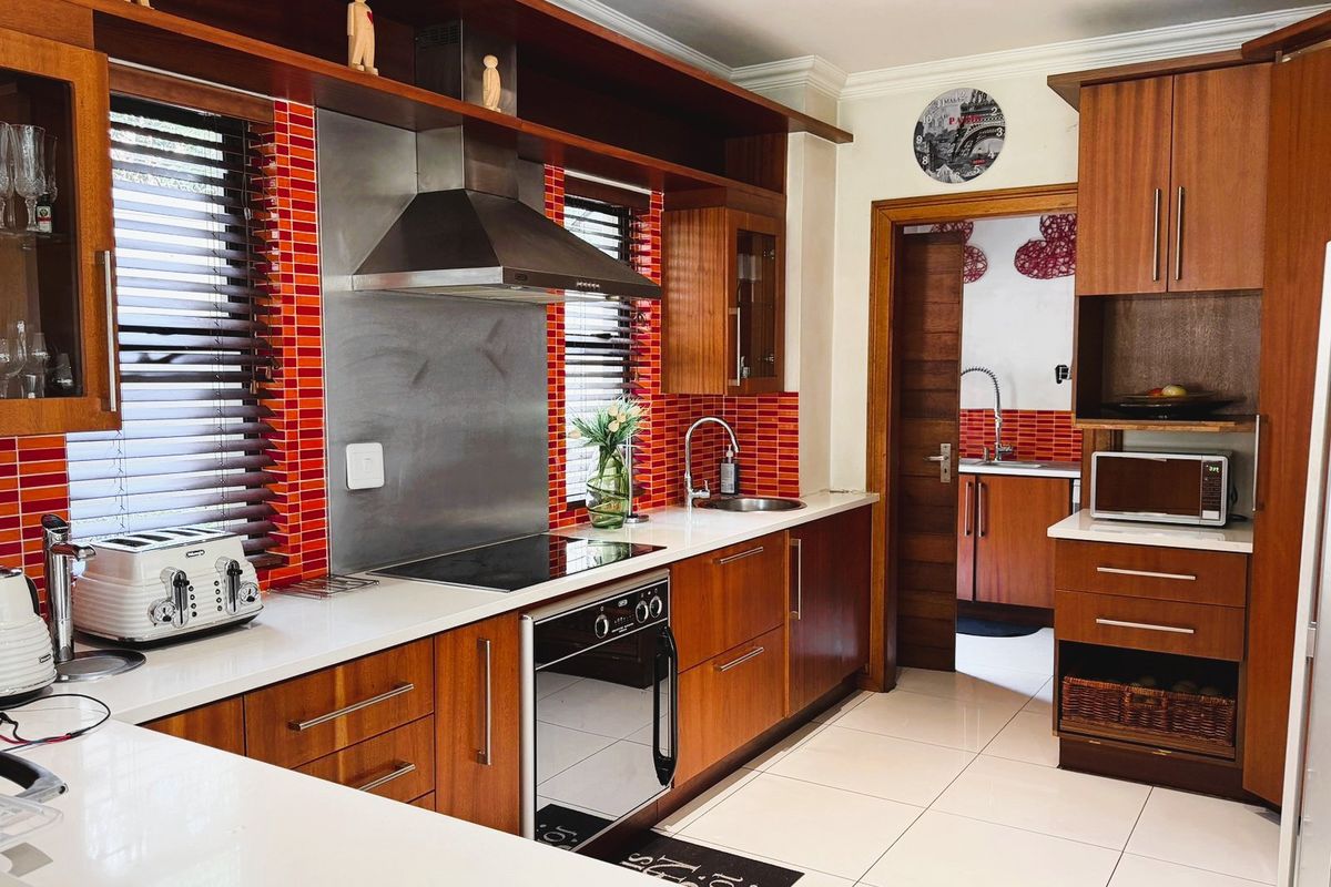 A well-maintained kitchen featuring warm wood cabinetry, stone countertops, a modern stainless steel extractor fan, and a vibrant red tiled splashback.