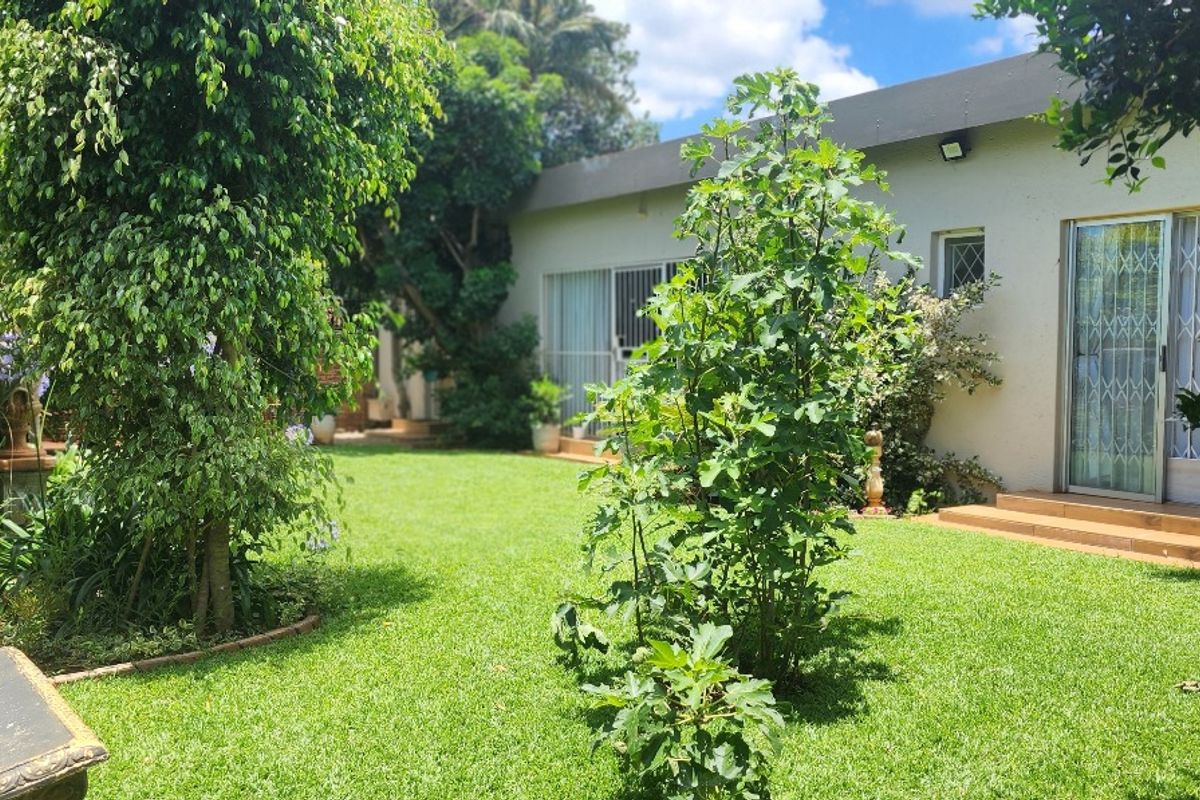 A view of the rear garden featuring a well-maintained lawn, mature trees, and access to the home via sliding glass doors with security gates.