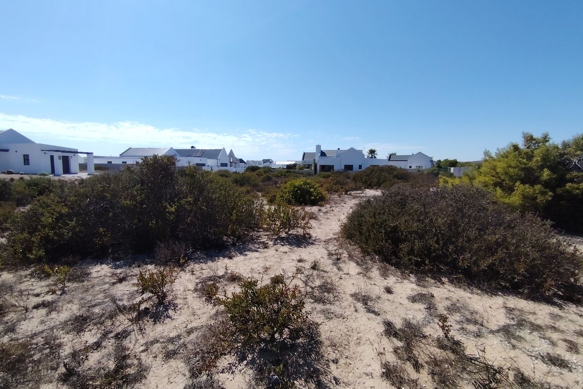 A view of a vacant residential plot featuring sandy ground and native coastal shrubs, with neighboring houses visible in the background.