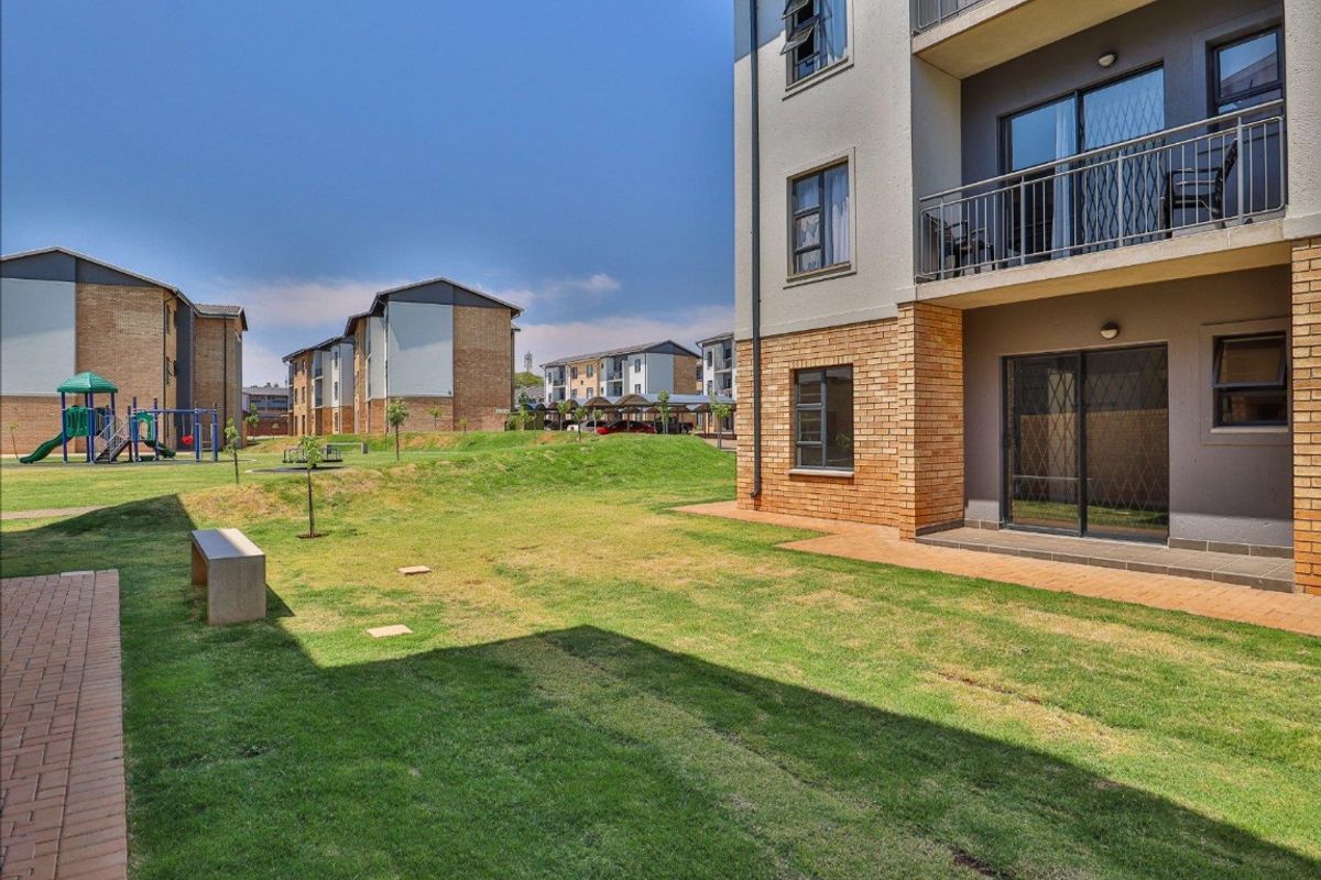A view of the apartment complex exterior featuring a shared garden area, paved patio, and brick facade with balcony access.