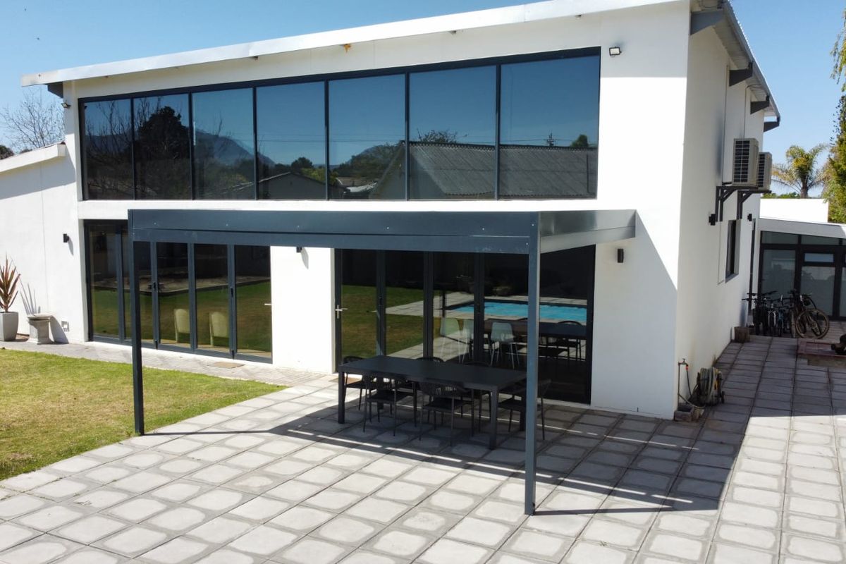 Rear view of a modern two-story home featuring a paved patio area with a covered pergola, large glass sliding doors, and expansive windows.