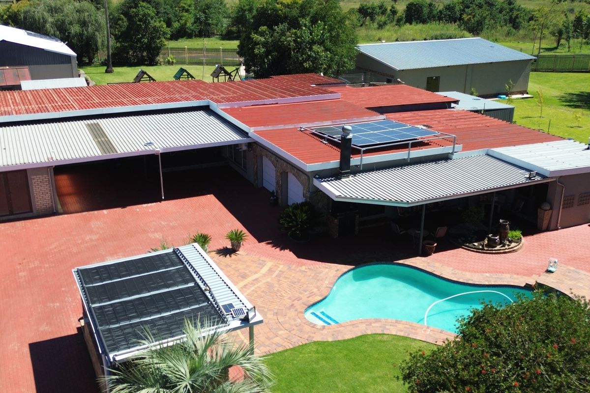 Aerial view of the property featuring a swimming pool with solar heating, extensive paved areas, and a large metal roof with integrated solar panels.