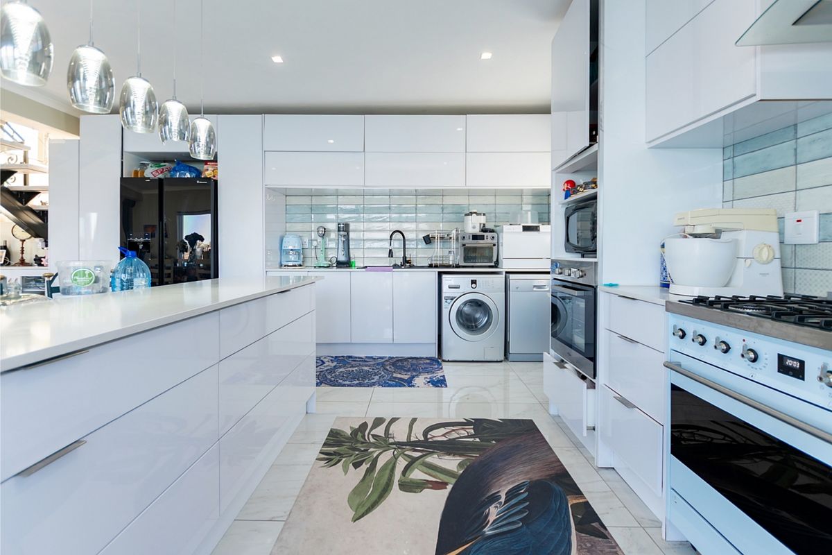Modern, high-gloss white kitchen featuring stone countertops, integrated appliances, a central island, and a stylish subway tile backsplash with recessed lighting.