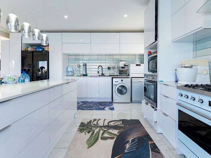 Modern, high-gloss white kitchen featuring stone countertops, integrated appliances, a central island, and a stylish subway tile backsplash with recessed lighting.