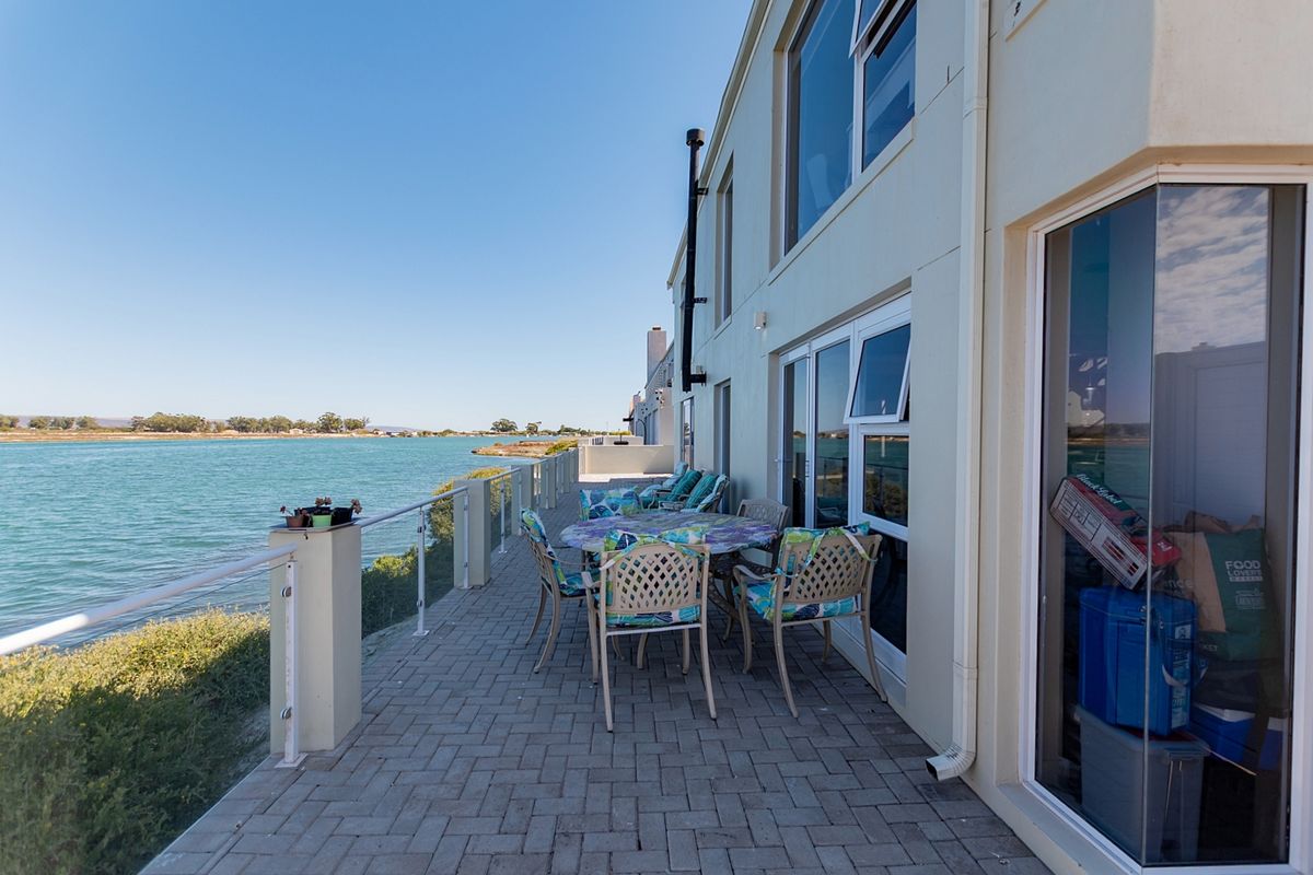 A spacious paved patio area featuring a glass balustrade, offering direct views of the adjacent waterway and access to the home via sliding doors.