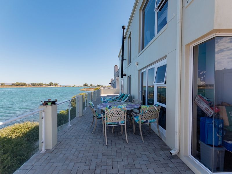 A spacious paved patio area featuring a glass balustrade, offering direct views of the adjacent waterway and access to the home via sliding doors.