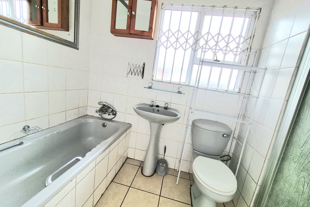 A dated bathroom featuring a bathtub, pedestal basin, and toilet, with tiled finishes and security bars on the window.