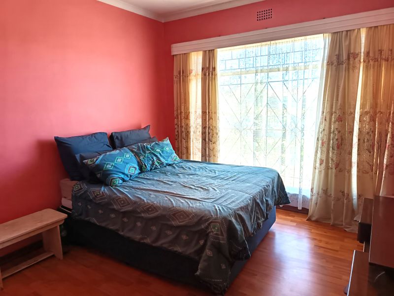 A bedroom featuring laminated timber flooring, a large window with security burglar bars, and a ventilation grille.