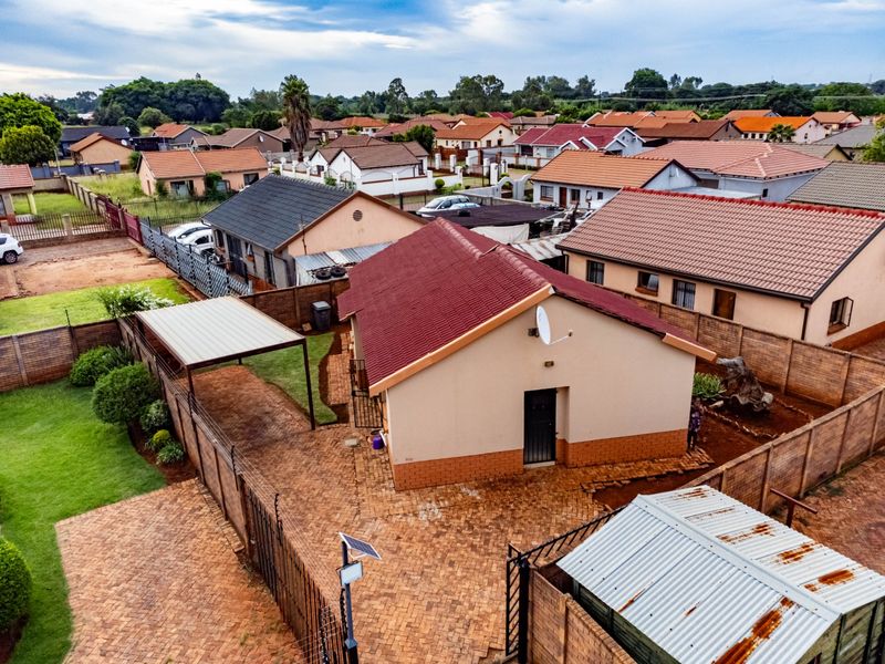 Aerial view of a residential property featuring a tiled roof house, paved driveway, carport, and perimeter walling in a suburban neighborhood setting.