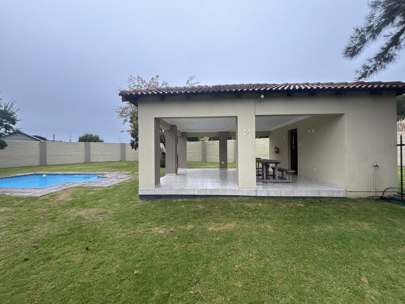 A covered outdoor patio area featuring tiled flooring and concrete pillars, overlooking a private garden and swimming pool.