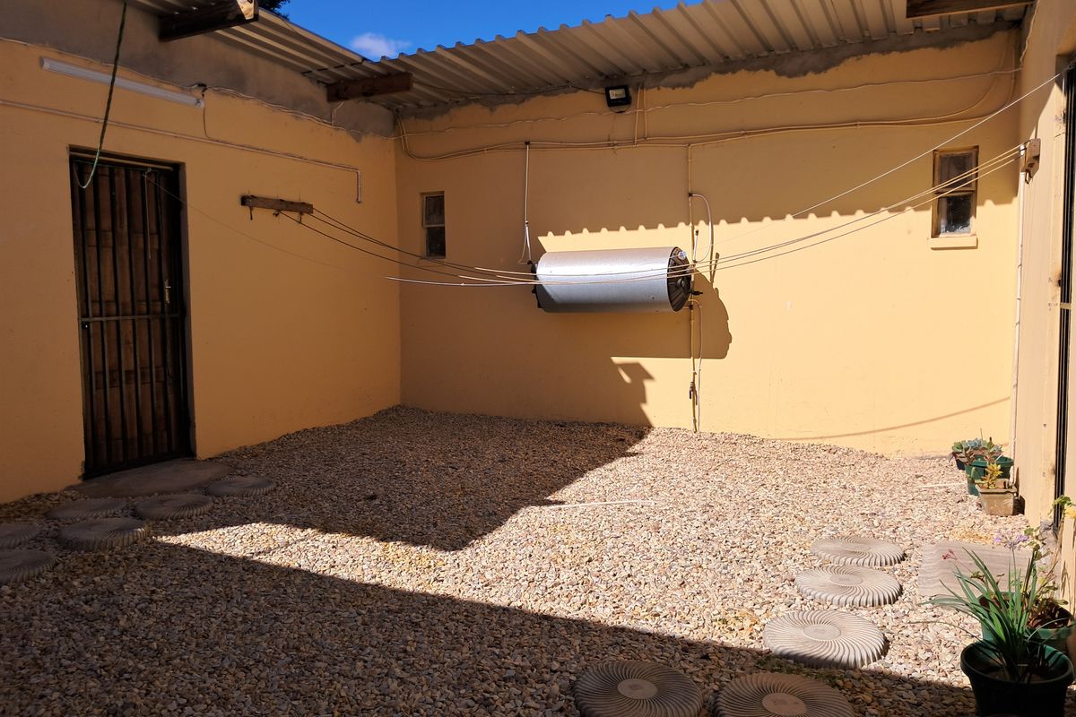 A rear courtyard area featuring a gravel surface, stepping stones, a security gate, and an externally mounted electric geyser.