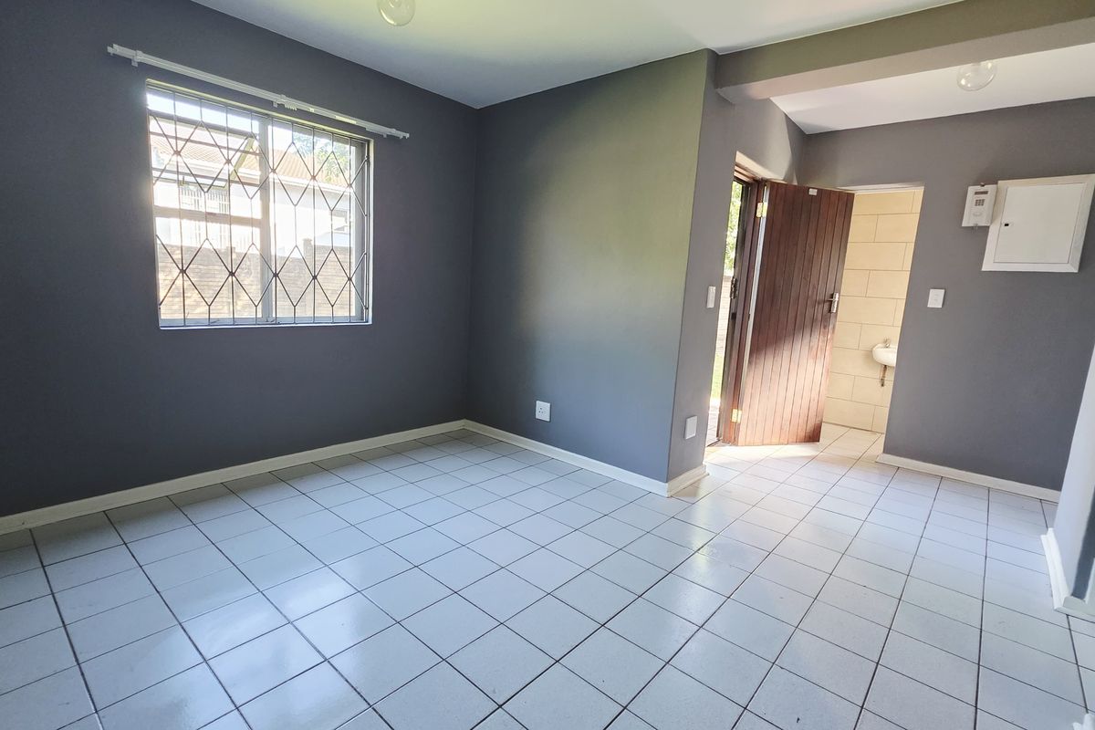 An empty living area featuring tiled flooring, grey painted walls, and a window with security bars, leading to an adjacent tiled space.