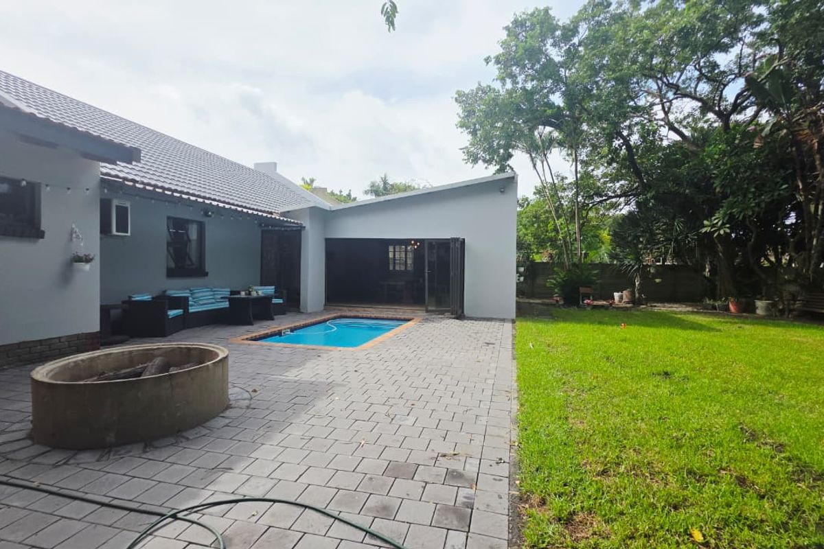 Rear exterior view featuring a paved patio, built-in boma fire pit, swimming pool, and a spacious garden area with mature trees.