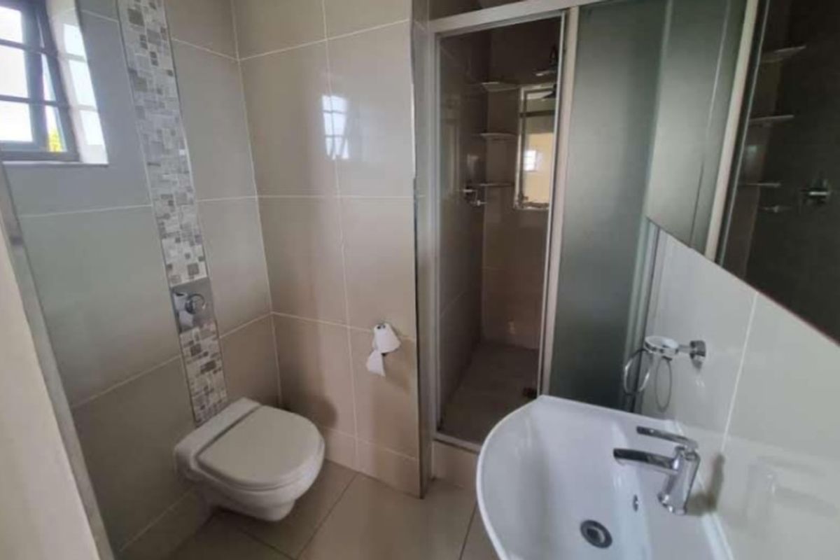 A clean, modern bathroom featuring a wall-mounted toilet, a glass-enclosed shower, and a pedestal basin with neutral wall and floor tiling.