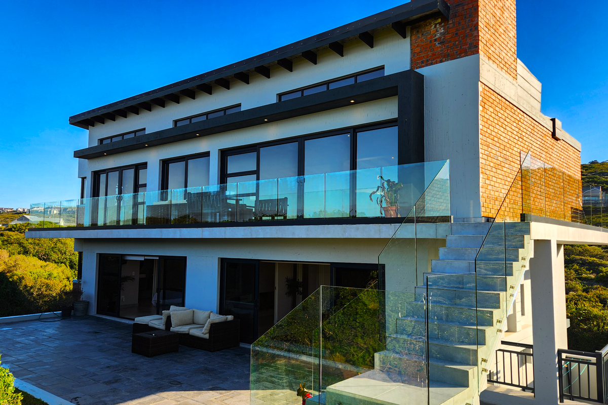 Modern multi-level rear elevation featuring a stone patio, glass balustrades, external staircase, and large sliding doors opening to the outdoor living space.