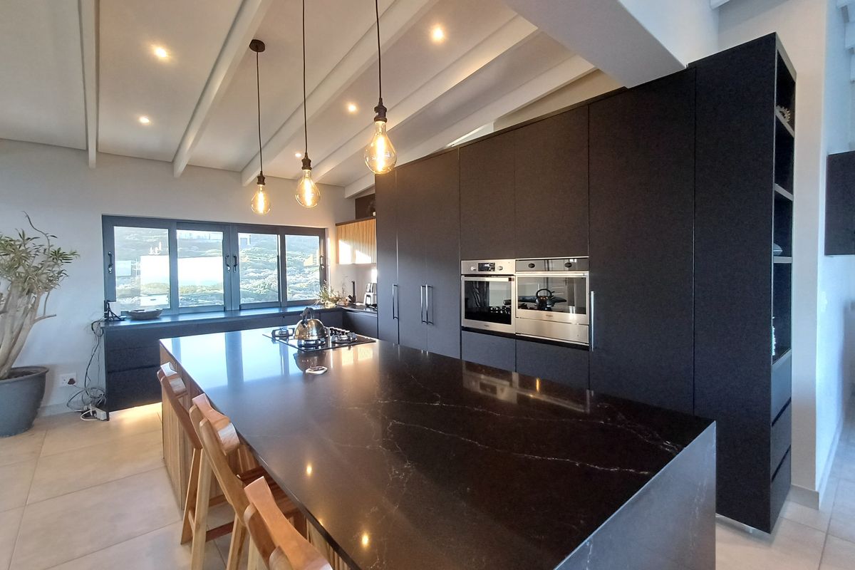 Modern kitchen featuring a large central island with stone countertops, integrated appliances, sleek dark cabinetry, and exposed ceiling beams with pendant lighting.