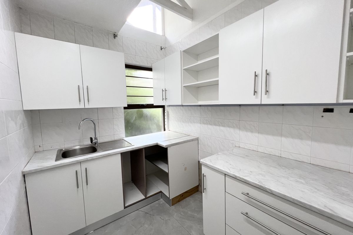 A functional kitchen featuring white cabinetry, marble-look countertops, and a stainless steel sink, with exposed roof trusses and natural light from a louvre window.