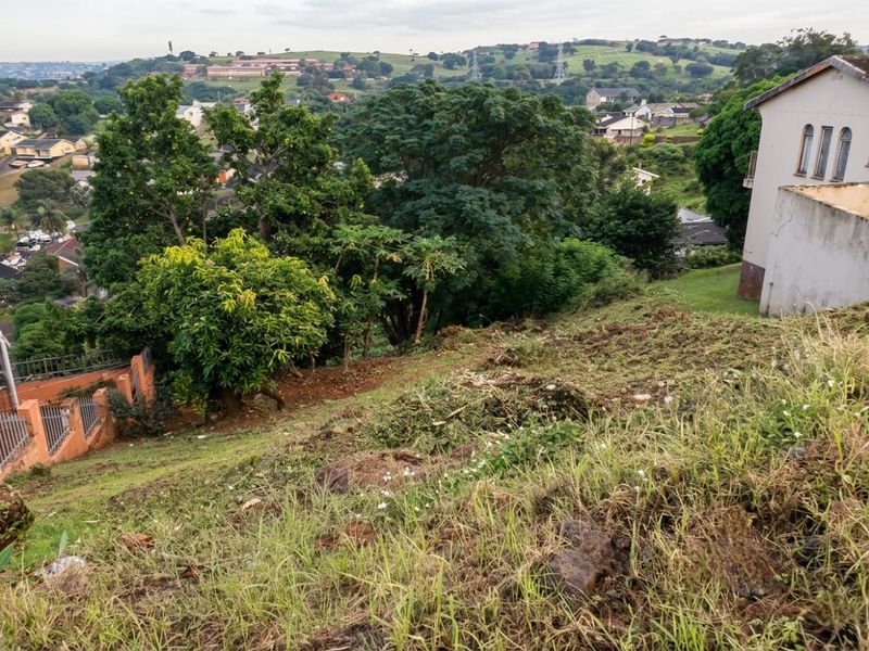 A view overlooking a sloped residential plot with mature trees and surrounding neighborhood scenery.