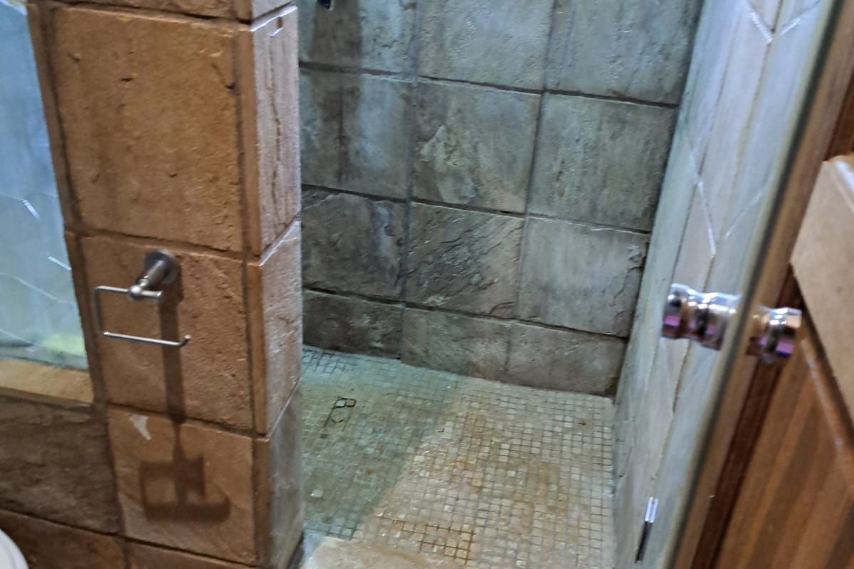 A bathroom shower enclosure featuring textured stone-look wall tiles and a mosaic tiled floor, requiring maintenance and updates to the tiling.