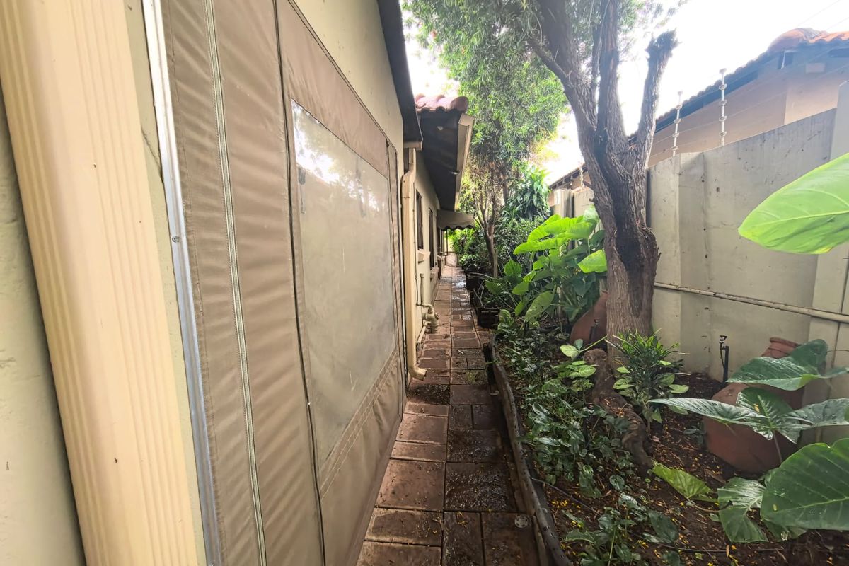 A narrow side garden path featuring paved flooring, boundary wall with electric fencing, and retractable canvas patio blinds attached to the building exterior.