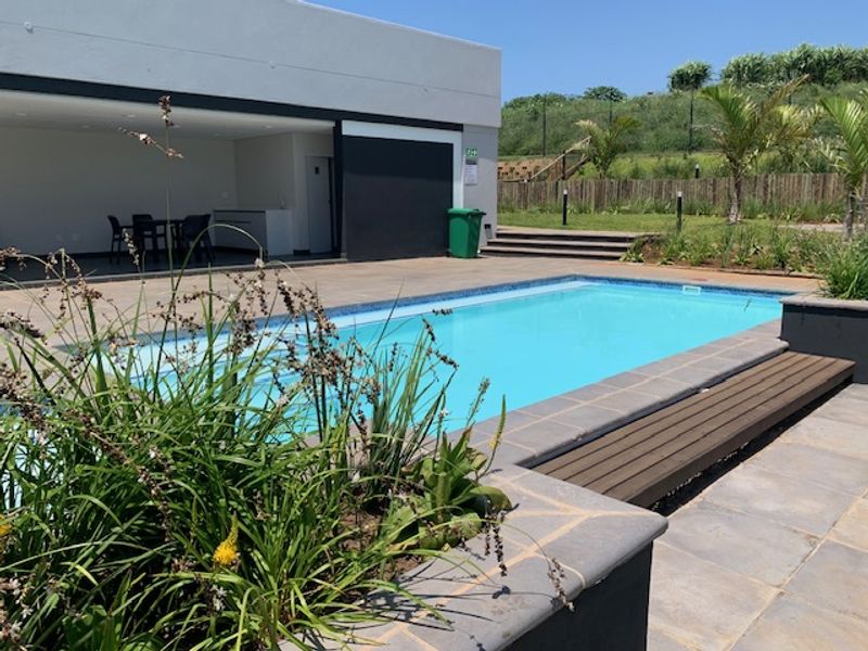 A modern outdoor pool area featuring a clear swimming pool, paved decking, and a covered patio space with a built-in service area.